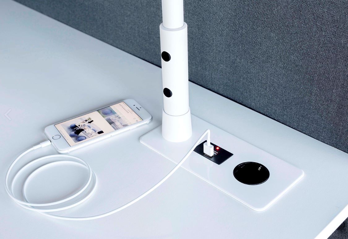 Detail van de witte lampvoet op een bureaublad, met stopcontact, oplaadplek en opladende telefoon