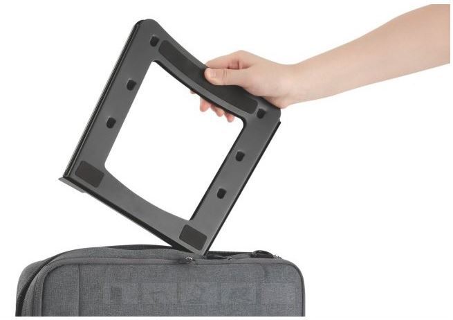 Een hand tilt de opgevouwen houder uit een tas, met het open frame en de rubberen randen in beeld