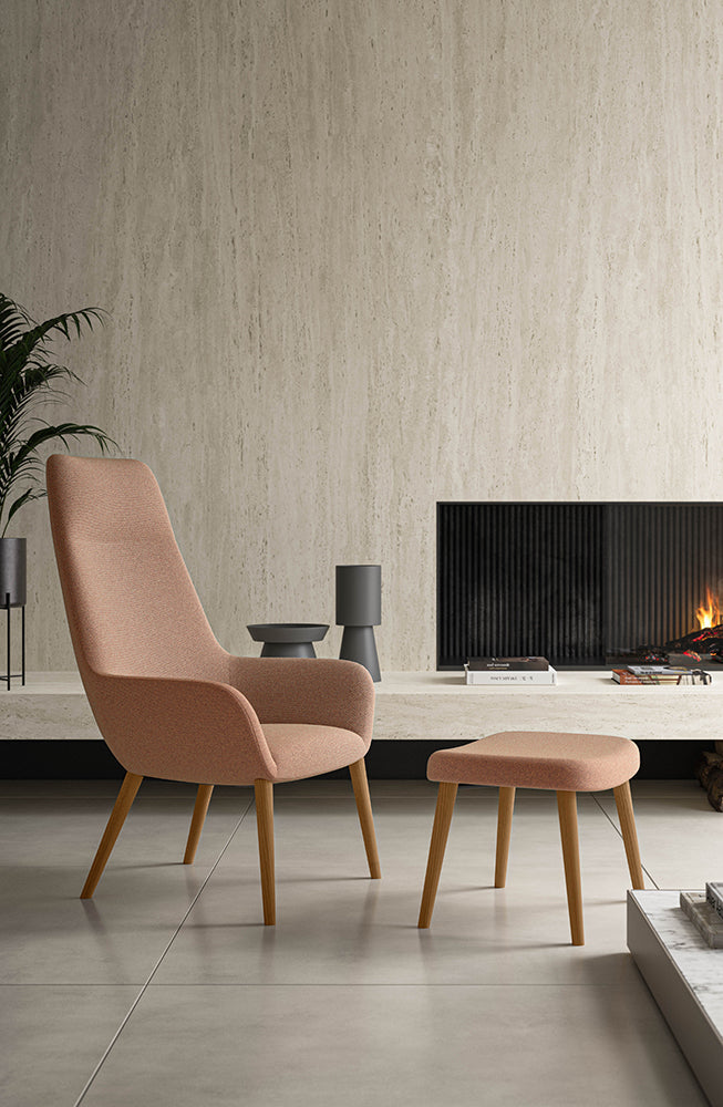 Sfeerbeeld van een zalmroze loungefauteuil met hoge rug en bijpassende voetenbank op houten poten bij een haard